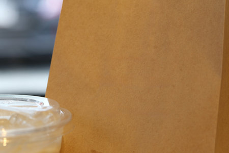 A close-up view of a blank brown paper bag alongside a plastic cup filled with ice, perfect for showcasing takeaway food and beverages in a cozy cafe environment.の写真素材