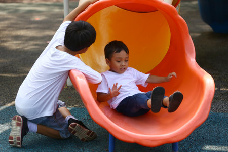 Two young boys are joyfully playing on a colorful slide at the playground, capturing the essence of childhood happiness and adventure in a vibrant outdoor setting.の写真素材