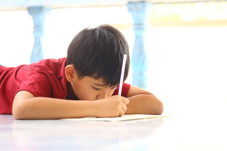 A young boy lies on the floor, deeply focused on writing, showcasing concentration and creativity in a bright outdoor setting filled with natural light.の写真素材