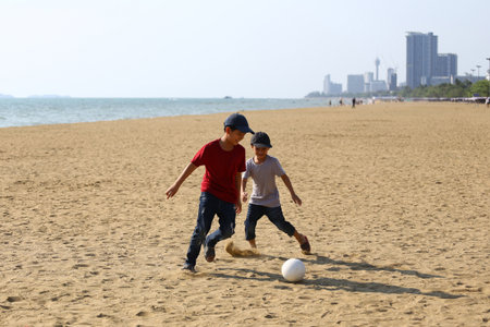 Two energetic young boys enjoy playing soccer on a sunny beach, surrounded by sand and a distant city skyline, creating a moment of joy and childhood adventure.の写真素材