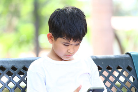 A young boy sitting on a bench is deeply focused on his smartphone screen in a sunny park, showcasing modern childhood and digital interaction amidst nature.の写真素材