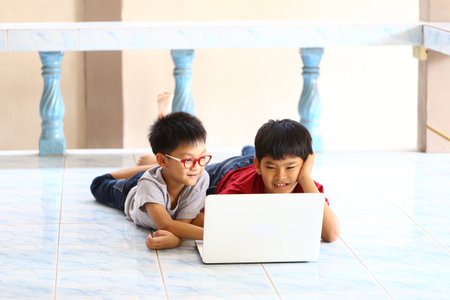 Two boys are captivated by a laptop screen, showcasing the joy of companionship and the power of technology in modern life. They lay on a tiled floor, immersed in their activity.の写真素材