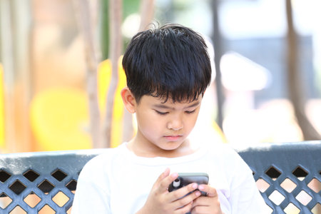 A young boy sits on a bench in a park, deeply focused on his smartphone, embodying a moment of concentration and engagement in a digital world.の写真素材