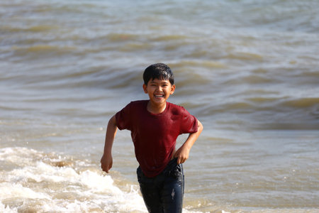 A cheerful young boy runs joyfully along the beach, splashing through the waves and capturing the essence of carefree childhood moments under the bright sun.の写真素材