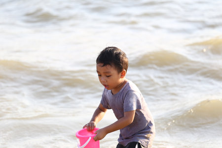 A young boy enjoys a sunny day at the beach, playing with a pink bucket in the water. The scene captures the joy of childhood and the beauty of nature.の写真素材