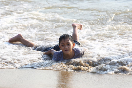A joyful young boy lies on the sandy shore, amidst gentle waves at the beach, capturing playful essence of childhood and the carefree spirit of summer adventures.の写真素材