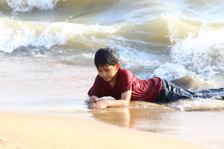 A young boy enjoys a fun-filled day at the beach, playing in the shallow water and experiencing the joy of nature amidst gentle waves and warm sunshine.の写真素材