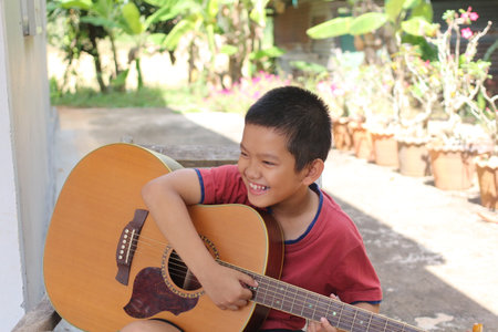 A young boy joyfully playing his acoustic guitar outdoors, surrounded by nature. His smile radiates happiness as he engages in music, capturing the essence of childhood and creativity.の写真素材