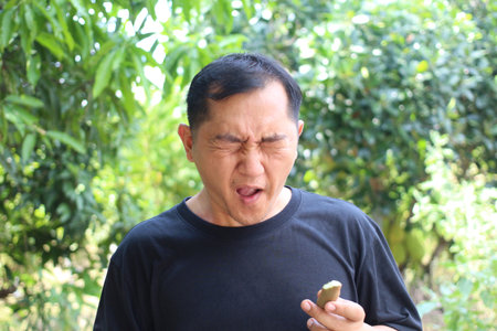 A man outdoors displays a disgusted expression while holding a fruit. Surrounded by lush greenery,...