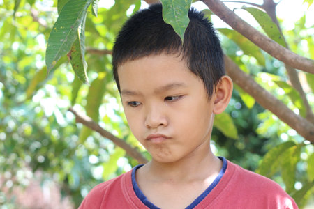 A young boy stands thoughtfully among lush green leaves, displaying a pensive expression. This...