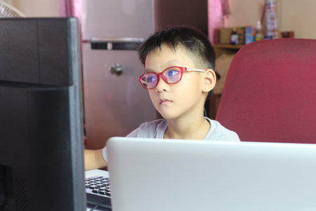 A young boy wearing glasses focuses intently on his computer in a home office setup, showcasing modern learning and technology experiences in a casual environment.の写真素材
