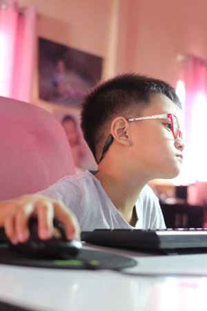 A young boy wearing glasses is focused on his computer at home. The warm lighting and relaxed...