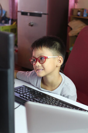 A young boy wearing red glasses enjoys his time at a computer. He is focused and smiling while...