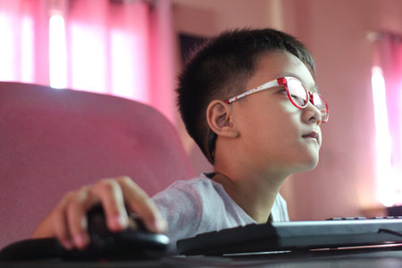 A young boy wearing glasses sits attentively at a computer, focused on the screen. The image captures concentration and the essence of childhood in a digital age.の写真素材