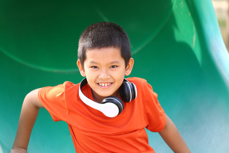 A joyful boy with headphones smiles brightly while playing on a green slide in the playground, embodying happiness and carefree childhood moments.の写真素材