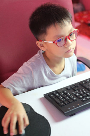 A young boy wearing glasses is attentively using a computer at home. The modern workspace reflects a learning and playful environment filled with technology.の写真素材