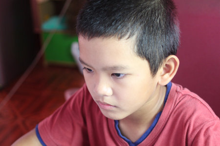 A close-up portrait of a focused young boy with short hair wearing a red shirt. The child's serious expression captures a moment of concentration in a casual indoor environment.の写真素材