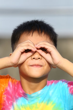 A cheerful child wearing a colorful tie-dye shirt makes a playful gesture with hands over eyes, showcasing joy and innocence in an outdoor setting.の写真素材