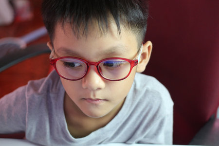 A portrait of a thoughtful boy wearing red glasses, lost in concentration. Captured in a casual setting, this image reflects innocence and curiosity.の写真素材