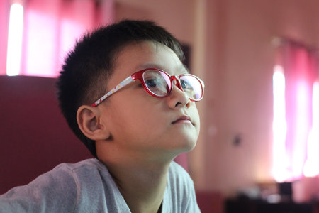 A young boy wearing red glasses gazes intently, immersed in thought. Bright indoor lighting enhances the serene atmosphere, showcasing curiosity and focus.の写真素材