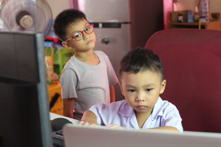 Two children explore technology at home, one focused on the computer. The scene captures the curiosity and playful engagement of youth in a nurturing environment.の写真素材