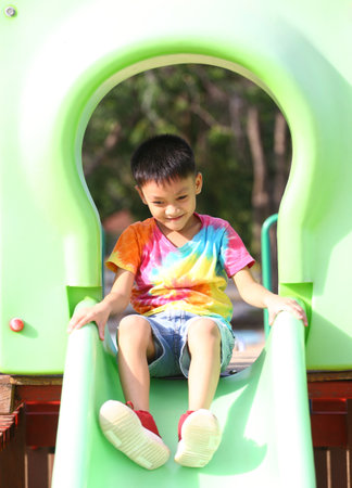 A young boy in a colorful shirt joyfully slides down a green playground slide, capturing the essence of childhood play and carefree adventure in an outdoor setting.の写真素材