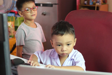 Two boys engage with a computer in a cozy home setting, showcasing curiosity and learning. One boy focuses intently while the other observes, capturing the essence of childhood exploration and technology.の写真素材