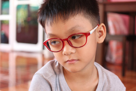 A thoughtful young boy in red glasses sits indoors, displaying a serious expression. This candid portrait captures the innocence and curiosity of youth in a warm home setting.の写真素材