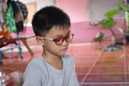 A young boy with glasses sits thoughtfully indoors, showcasing innocence and curiosity. The relaxed setting captures a moment of contemplation in childhood.の写真素材