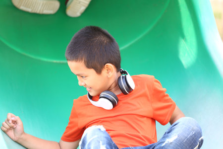 A joyful boy wearing headphones slides down a green playground slide, showcasing the essence of childhood fun and carefree moments in the outdoors.の写真素材