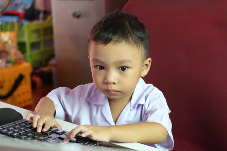 A focused young child sitting at a computer desk, engaging with a keyboard. This image captures the essence of childhood curiosity and early learning in a casual indoor setting.の写真素材