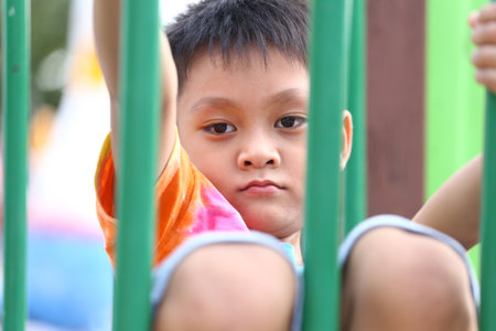 A young child sits thoughtfully on playground bars, showcasing a vibrant shirt. The image captures a moment of contemplation against an outdoor backdrop.の写真素材