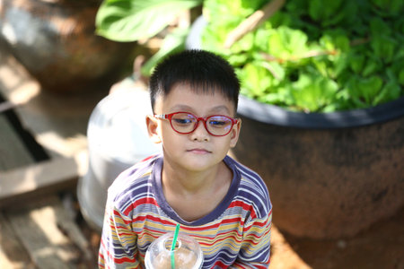 A young boy with glasses enjoys a refreshing drink in a vibrant outdoor setting, showcasing a moment of childhood joy surrounded by greenery.の写真素材
