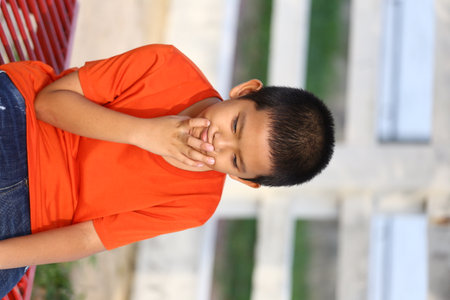 A young boy sits outdoors in a bright orange shirt, lost in thought. His casual pose and natural setting capture the essence of childhood wonder and innocence.の写真素材