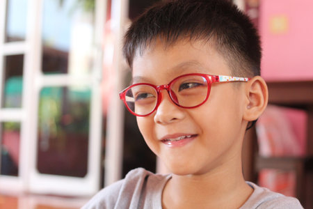 A cheerful boy with red glasses smiles brightly in a warm indoor setting. This joyful portrait captures the essence of childhood and happiness.の写真素材