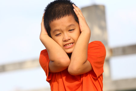 A young boy in an orange shirt covers his ears with a distressed expression in an outdoor setting, capturing a moment of vulnerability and emotion.の写真素材