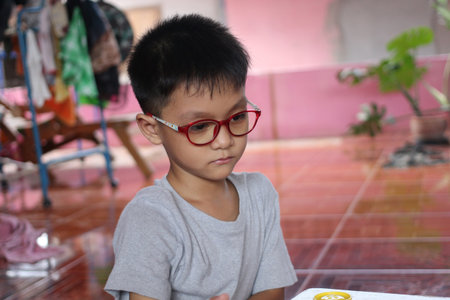A thoughtful young boy wearing red glasses sits indoors, focused and serene. The warm atmosphere highlights his emotions in a casual setting.の写真素材