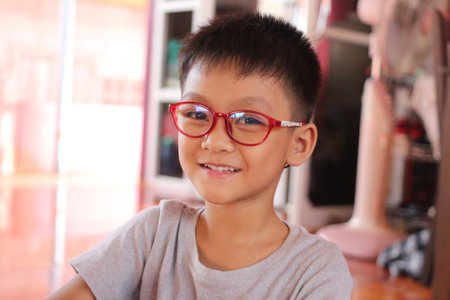 A cheerful young boy with red glasses smiles warmly in a cozy indoor setting. This portrait captures the joy and playfulness of youth in a casual atmosphere.の写真素材