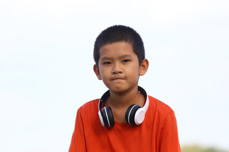 A young boy wearing headphones sits outdoors, enjoying music. His casual orange shirt highlights a cheerful and carefree expression, reflecting youth and happiness.の写真素材