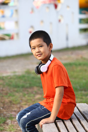 A young boy with headphones sits casually on a wooden bench in a park, enjoying the outdoors. His orange shirt contrasts with the natural setting, capturing a moment of joy.の写真素材