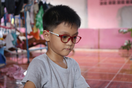 Young boy wearing red glasses sits thoughtfully in a vibrant indoor space, showcasing a moment of concentration and youthful curiosity amidst a lively backdrop.の写真素材