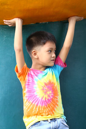 A joyful young boy in a colorful tie-dye shirt hangs playfully under a bright structure, radiating energy and innocence in a vibrant outdoor setting.の写真素材