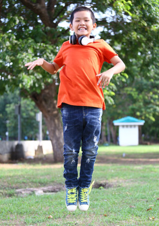 A cheerful boy demonstrates pure joy while jumping in a park. His bright orange shirt and headphones reflect a fun, active lifestyle amidst natural surroundings.の写真素材