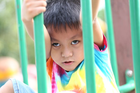 A young child demonstrates curiosity while playing on a playground, grasping the bars with an inquisitive expression. The vibrant colors of the t-shirt add to the joyful atmosphere.の写真素材