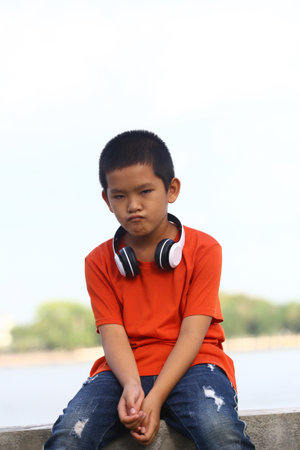 A young boy with headphones sits outdoors, displaying a thoughtful expression. Dressed casually in an orange shirt, he embodies the essence of youth and technology in a vibrant setting.の写真素材