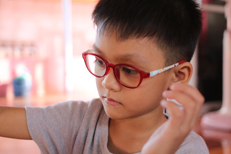 A young boy wearing red glasses shows concentration while engaging in an activity. The close-up captures his thoughtful expression, reflecting curiosity and innocence in a bright, playful environment.の写真素材