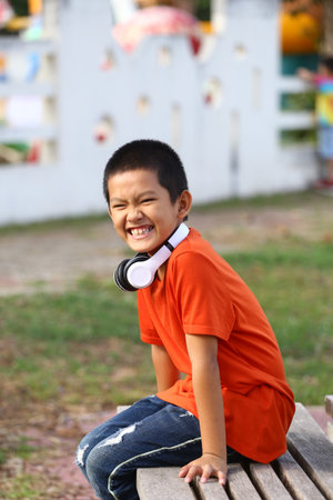 A cheerful boy wearing headphones smiles brightly while sitting on a bench in an outdoor setting. His relaxed pose captures the essence of childhood joy.の写真素材
