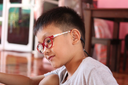 A young boy wearing red glasses sits indoors, conveying a sense of focus and creativity. The colorful background adds warmth to this candid moment.の写真素材