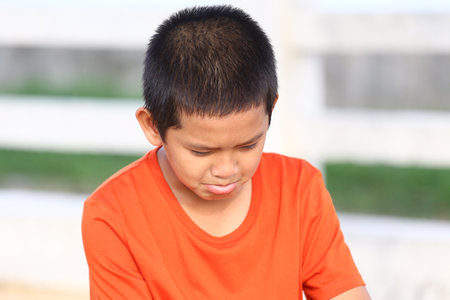 A young boy wearing a bright orange shirt displays a sad expression outdoors. His downcast gaze and frown evoke a sense of contemplation and emotion.の写真素材