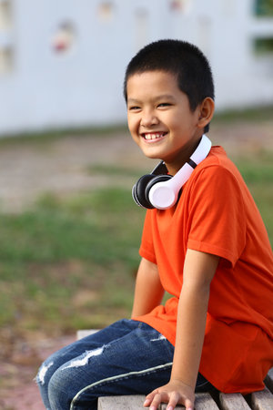 A joyful boy wearing headphones sits outside with a big smile. His orange shirt and relaxed posture highlight carefree childhood moments, perfect for music enjoyment.の写真素材
